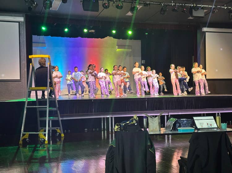A group of students on stage in matching flare pants and white tops dancing a routine dance together. Background lighting has rainbow colours vertifclly down.