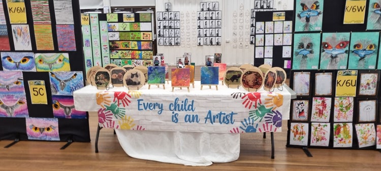 A photo of alot of children bright artwork on black stands and on the walls, with a table with a white table cloth saying