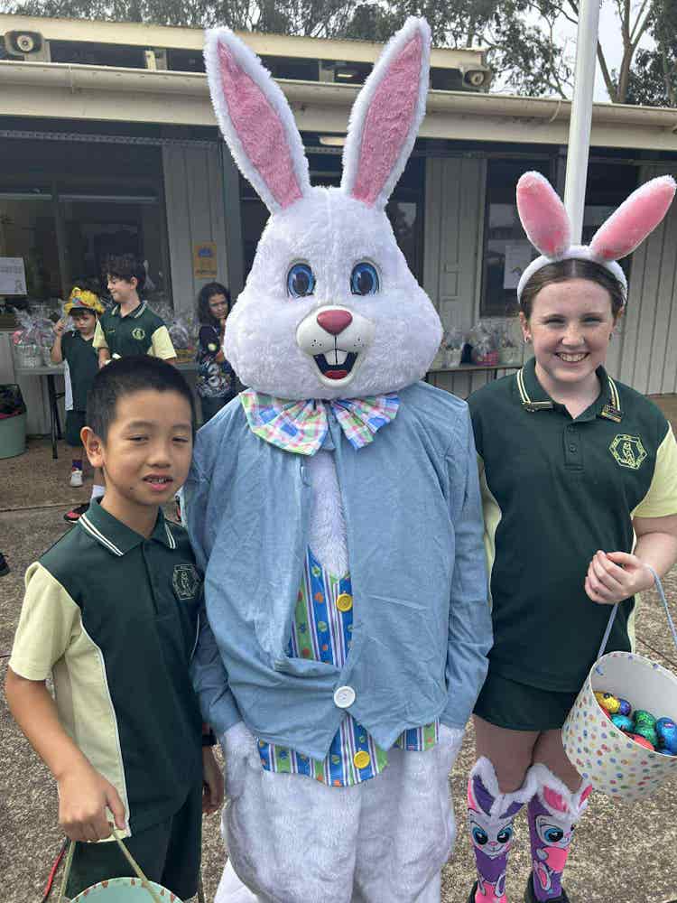 A photo of who students standing either side of a person dressed as the easter bunny. Celebrating our easter hat parade