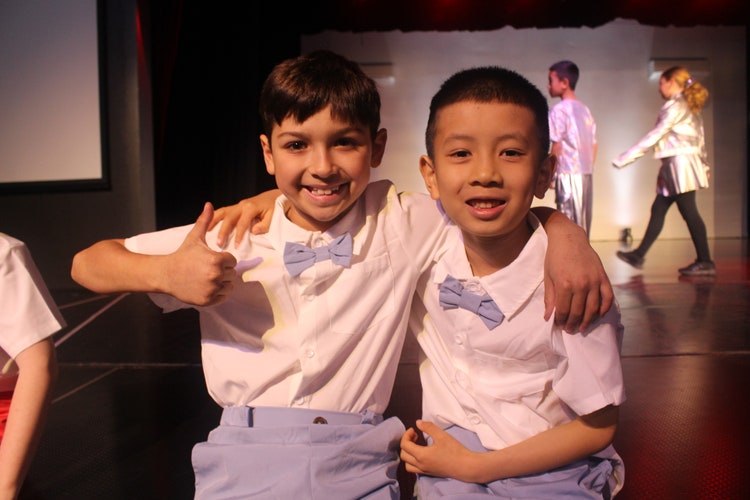 A photo of two students sitting on the edge of the stage dressed in blue bowties, one boy has his arm around the other student with a thumbs up.