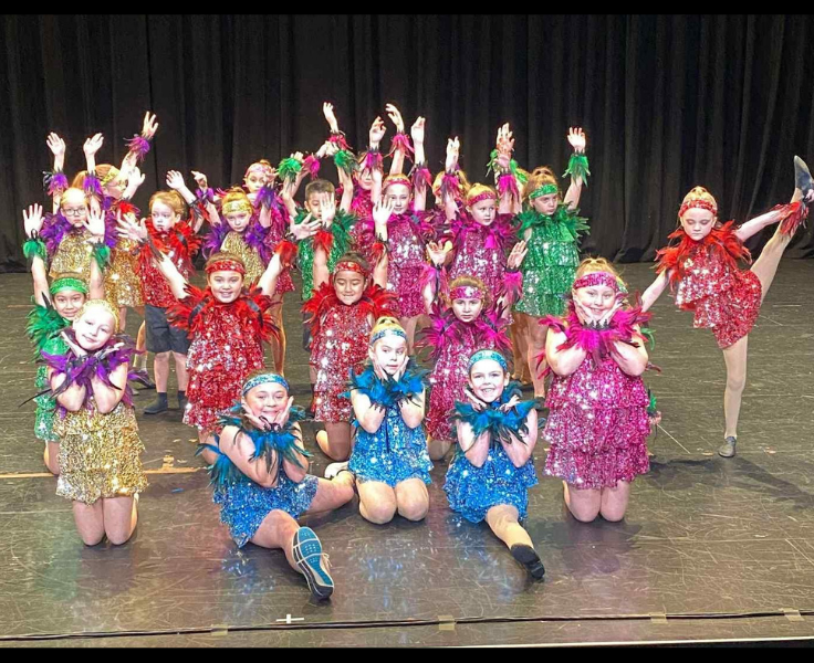 A group of around 20 students dressed in brightly and shinny dance costumes on stage after performing a dance.