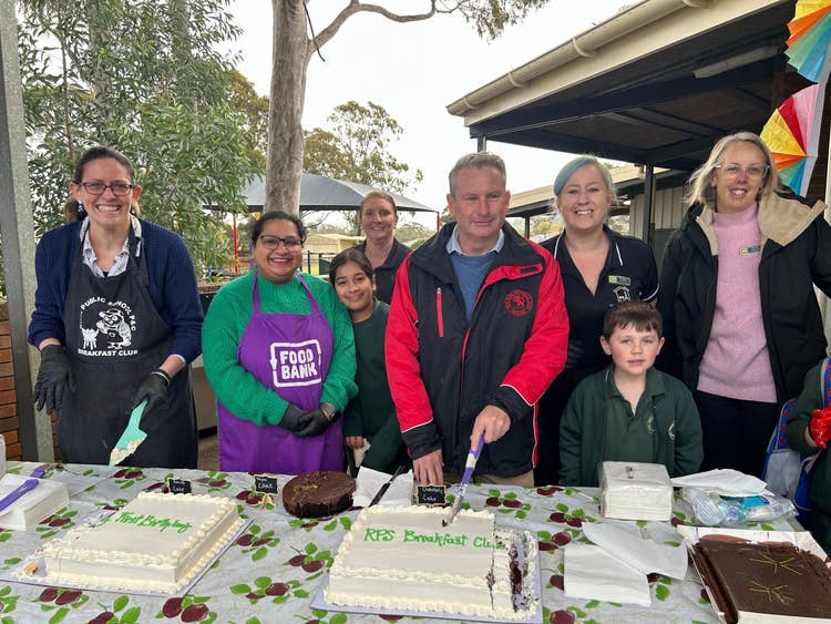8 people standing behind a table cutting a big white cake celebrating breakfast clubs first birthday.